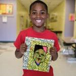 A young student holds up a piece of student art and smiles for a photo